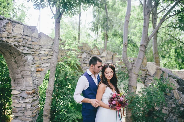 Happy groom in a dark blue suit hugs a beautiful bride in a white wedding dress with pink and purple bouquet of flowers outdoors on the background of a stone arch — Stock Photo, Image