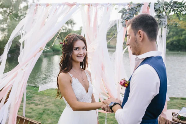 Noivo feliz em um terno azul escuro segurando a mão de sua bela noiva sorridente em um vestido de casamento branco ao ar livre sobre as decorações de fundo das fitas e lago — Fotografia de Stock