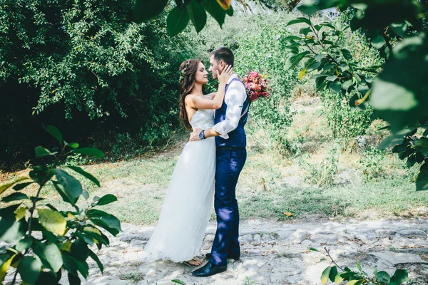 Felice sposo in un abito blu scuro abbraccia una bella sposa in un abito da sposa bianco con bouquet rosa e viola di fiori all'aperto — Foto Stock