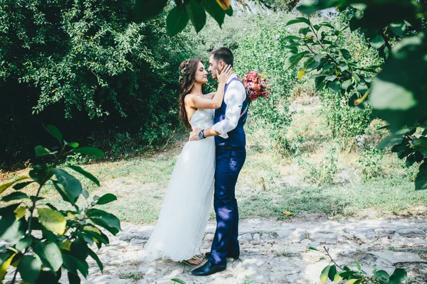 Happy groom in a dark blue suit hugs a beautiful bride in a white wedding dress with pink and purple bouquet of flowers outdoors — Stock Photo, Image