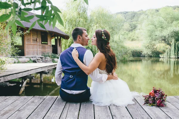Noivo feliz em um terno azul escuro abraça uma noiva bonita em um vestido de noiva branco e uma grinalda de flores ao ar livre em uma ponte de madeira no fundo do lago — Fotografia de Stock