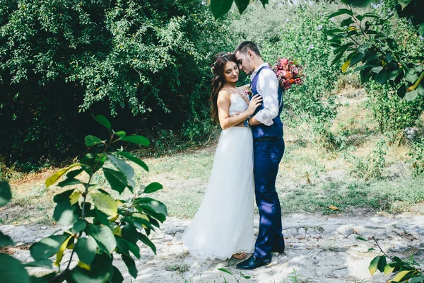 Happy groom in a dark blue suit hugs a beautiful bride in a white wedding dress with pink and purple bouquet of flowers outdoors — Stock Photo, Image