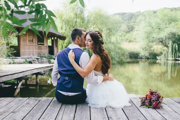 Noivo feliz em um terno azul escuro beija uma noiva bonita em um vestido de noiva branco e uma grinalda de flores ao ar livre em uma ponte de madeira — Fotografia de Stock