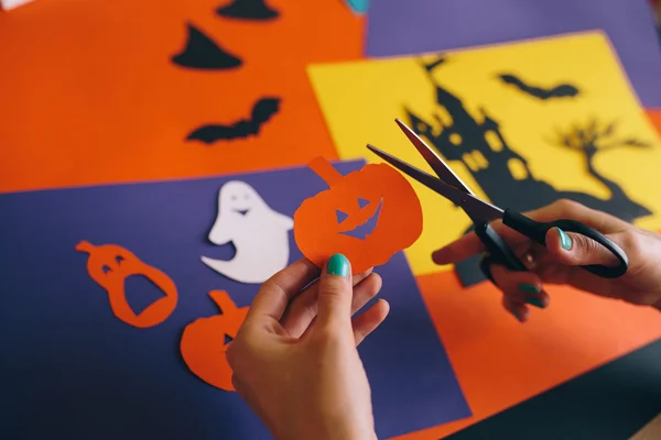 Primer plano de las manos femeninas con tijeras papel cortado calabaza naranja sobre el fondo composición de Halloween. Castillo, fantasmas, murciélagos, calabazas se cortan de papel de colores. Decoraciones caseras para Halloween —  Fotos de Stock