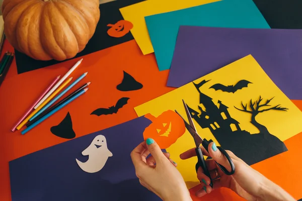 Fermer les mains féminines avec des ciseaux couper citrouille en papier sur le fond composition Halloween. Château, fantômes, citrouilles, chauves-souris et autres décorations effrayantes faites à la main . — Photo