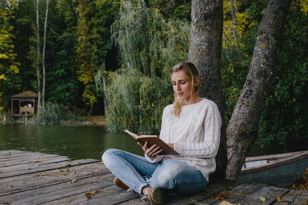 Jovem mulher loira em uma camisola branca e jeans azul lê um livro enquanto sentado em um cais de madeira no fundo da floresta de outono e lago — Fotografia de Stock