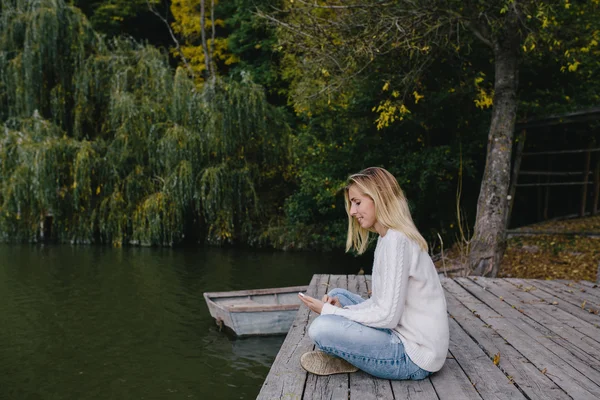 Mujer rubia joven en un suéter blanco y vaqueros azules sentados en un muelle de madera en el fondo del bosque de otoño, lago y el uso de teléfono inteligente. mujer que envía un mensaje de texto desde su teléfono celular al aire libre — Foto de Stock