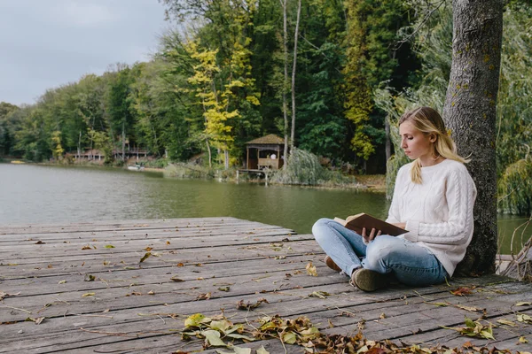 Jovem mulher loira em uma camisola branca e jeans azul lê um livro enquanto sentado em um cais de madeira no fundo da floresta de outono e lago — Fotografia de Stock