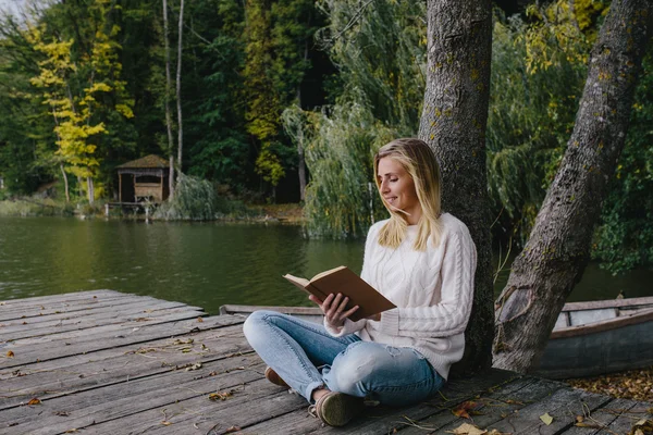 Jovem mulher loira em uma camisola branca e jeans azul lê um livro enquanto sentado em um cais de madeira no fundo da floresta de outono e lago — Fotografia de Stock