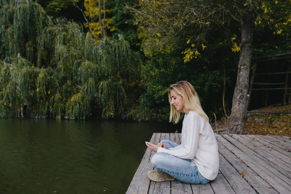 Jovem mulher loira em uma camisola branca e jeans azul sentado em um cais de madeira no fundo da floresta de outono, lago e usando telefone inteligente. mulher enviando uma mensagem de texto de seu telefone celular ao ar livre — Fotografia de Stock