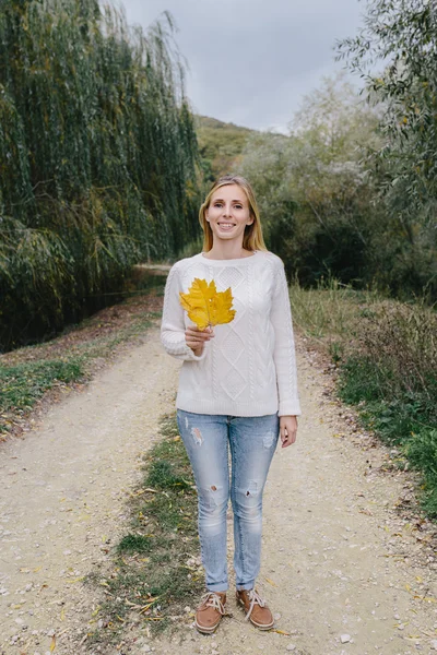Jeune femme souriante en pull blanc et jean bleu debout sur une route de campagne et tenant une feuille jaune tombée — Photo