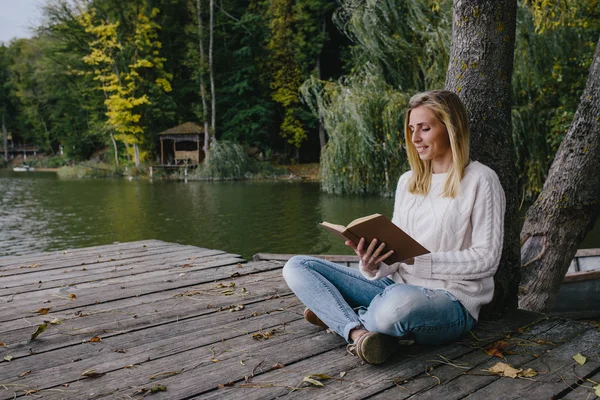 Młoda blondynka w białym swetrze i niebieskich dżinsach czyta książkę siedząc na drewnianym molo na tle jesiennego lasu i jeziora — Zdjęcie stockowe