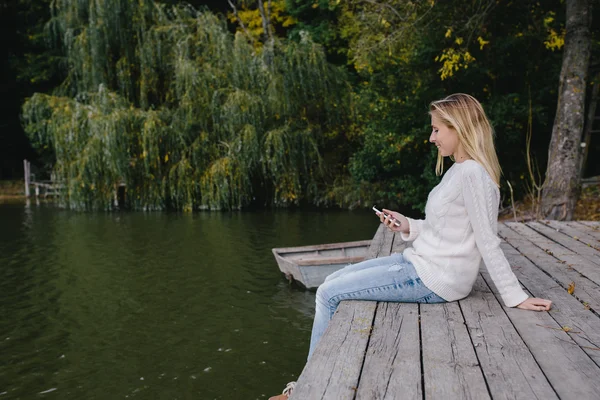 Junge blonde Frau in weißem Pullover und blauer Jeans, die auf einem Holzsteg vor dem Hintergrund von Herbstwald, See und Smartphone sitzt. Frau schickt SMS vom Handy ins Freie — Stockfoto