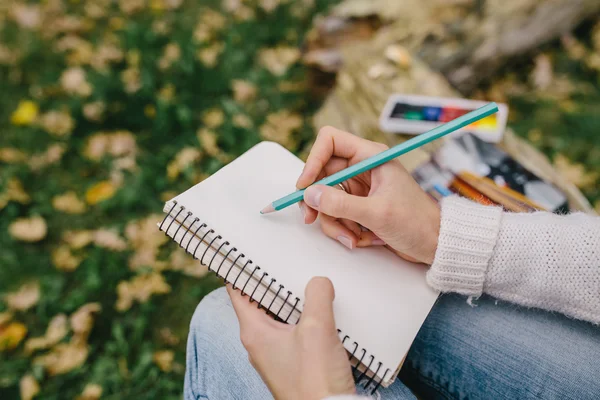 cropped image of a female hand drawing colored pencil in a notebook on the background of grass and fallen leaves. close up woman writing in a notebook outdoors