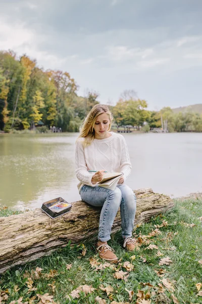 Pretty young woman in white sweater and blue jeans draws in a notebook with colored pencils sitting on trunk of a dry tree on a background of lake