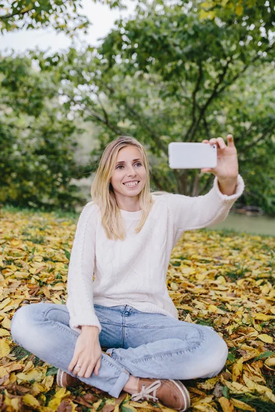Mujer joven bastante sonriente usando un teléfono móvil para tomar fotos al aire libre. mujer en un suéter blanco y pantalones vaqueros azules hace selfie sentado en la hierba y hojas caídas en el parque — Foto de Stock