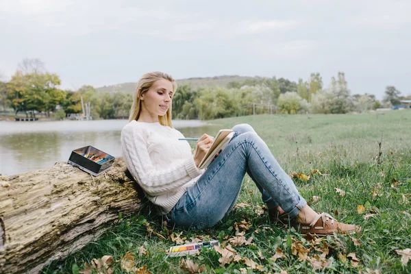 Muito sorrindo jovem mulher em camisola branca e jeans azul desenha em um caderno com lápis de cor sentado na grama e inclinado para o tronco de uma árvore seca em um fundo de lago — Fotografia de Stock