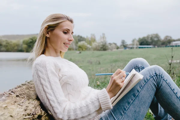 Muito sorrindo jovem mulher em camisola branca e jeans azul desenha em um caderno com lápis de cor sentado na grama e inclinado para o tronco de uma árvore seca em um fundo de lago — Fotografia de Stock