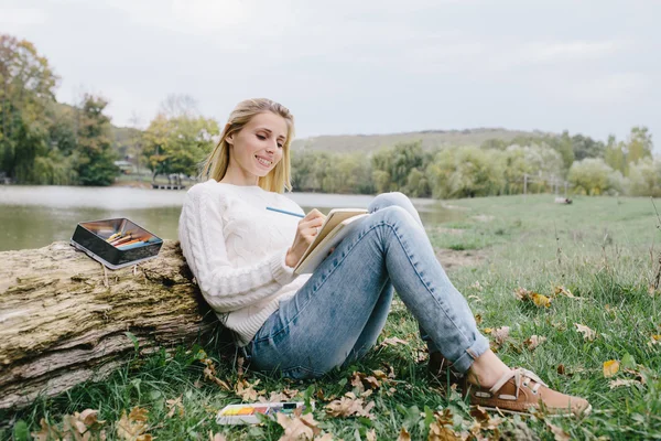 Muito sorrindo jovem mulher em camisola branca e jeans azul desenha em um caderno com lápis de cor sentado na grama e inclinado para o tronco de uma árvore seca em um fundo de lago — Fotografia de Stock