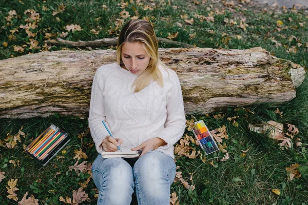 Mulher muito jovem em camisola branca e jeans azul desenha em um caderno com lápis de cor sentado na grama e inclinado para o tronco de uma árvore seca — Fotografia de Stock