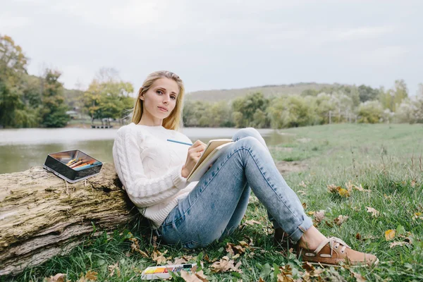 Ładna młoda kobieta w białym swetrze i niebieskie dżinsy rysuje w Notatniku z kolorowych ołówków siedzących na trawie i pochylił się w bagażniku suchego drzewa na tle jeziora — Zdjęcie stockowe