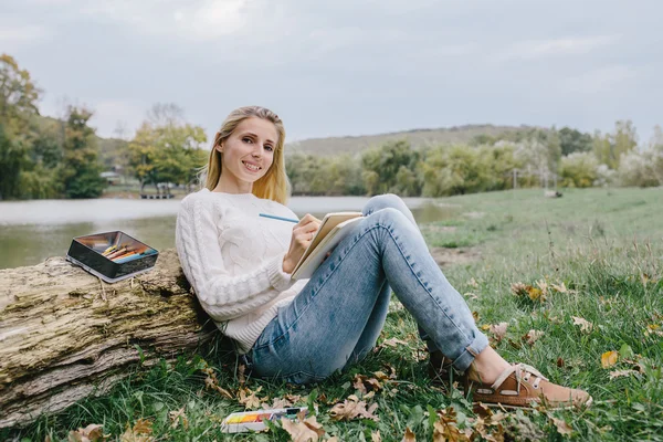 Pretty smiling young woman in white sweater and blue jeans draws in a notebook with colored pencils sitting on the grass and leaned into the trunk of a dry tree on a background of lake