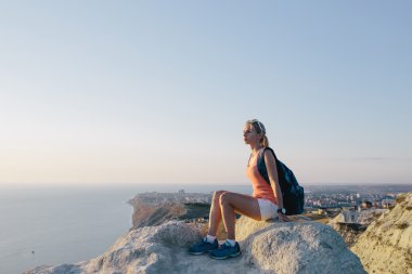 güneş gözlüğü ile aktif genç kadın turist bir dağın tepesinde oturan ve ileri deniz, gökyüzü ve şehir arka planda bakıyor.