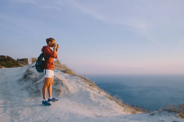 Giovane donna bionda prende la foto sulla macchina fotografica retrò su uno sfondo di cime di montagna, scogliere e il mare — Foto Stock