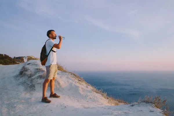 Young man with backpack drinking water on top of the mountain in the background of the sea — 图库照片