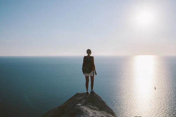 Mujer joven excursionista con mochila de pie en el acantilado y mirando hacia adelante en el fondo del mar, el cielo. mujer turista en la cima de una montaña disfrutando de la vista —  Fotos de Stock