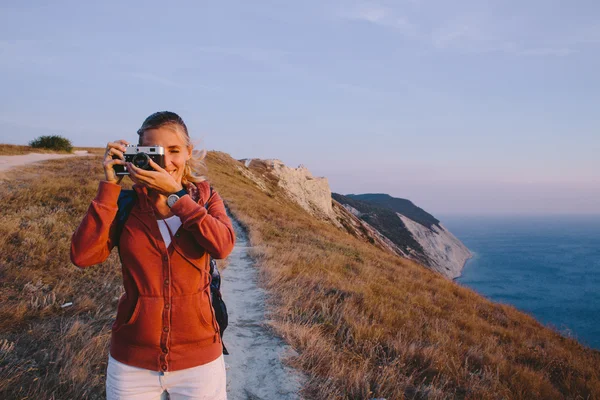Jovem loira tira a foto na câmera retro em um fundo de picos de montanha, falésias e o mar — Fotografia de Stock