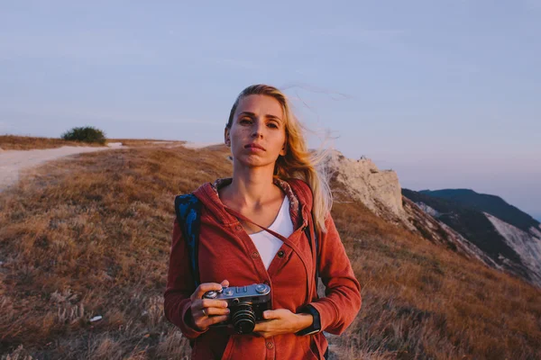 Portrait of attractive young blonde woman tourist standing with an retro camera in hands on a background of mountain peaks, cliffs and the sea — Stock Photo, Image