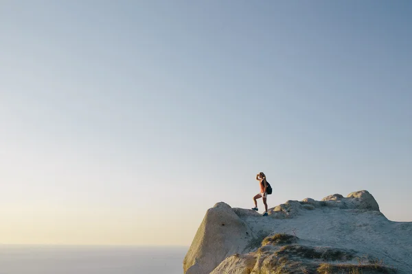 Young woman hiker with backpack standing on cliff and looking forward on the background of the sea, sky and city. lady tourist on top of a mountain enjoying view — 图库照片
