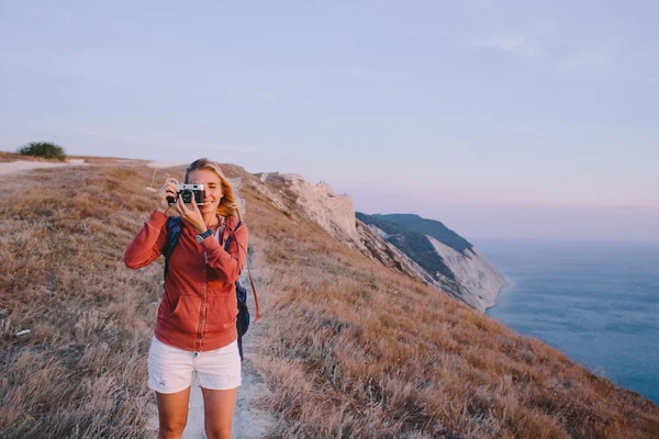 Young blonde woman takes the photo on the retro camera on a background of mountain peaks, cliffs and the sea — Stock Photo, Image