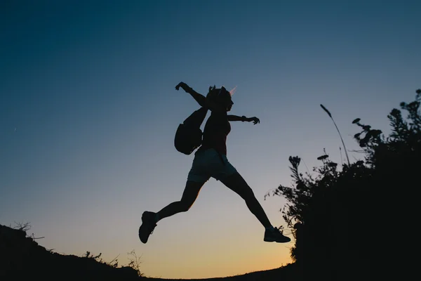 Silhueta jovem turista com mochila pulando através da lacuna entre colina. Mulher pulando sobre uma ravina — Fotografia de Stock
