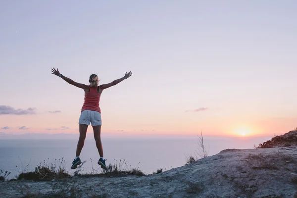 Feliz jovem caminhante pulando na montanha e olhando para a frente no mar e céu pôr do sol com as mãos levantadas — Fotografia de Stock