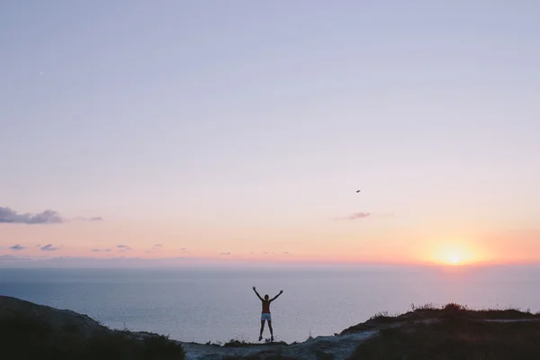 Jovem caminhante feliz em pé em uma cavidade entre duas colinas e olhando para a frente no mar e céu pôr do sol com as mãos levantadas — Fotografia de Stock