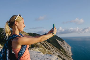 Young woman hiker with backpack use mobile phone taking photo on the background mountain peak cliff and sea