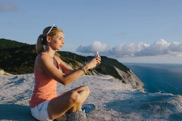 Mujer joven haciendo yoga posición de loto uso de teléfono móvil tomar fotos en el acantilado pico de montaña de fondo — Foto de Stock