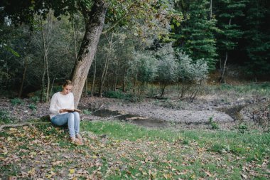 Beyaz kazak ve kot genç kadın parkta bir ağacın altında bir kitap okur
