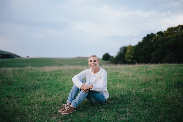 Szczęśliwa młoda kobieta w biały sweter i niebieskie dżinsy relaksujący na trawie w łące. kobieta korzystających życia na zewnątrz. — Zdjęcie stockowe