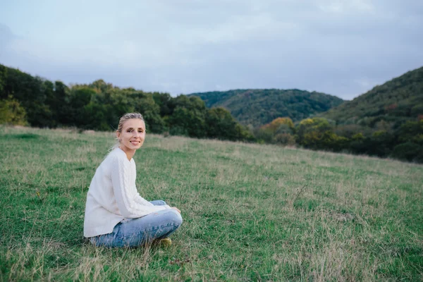Heureuse jeune femme en pull blanc et jean bleu relaxant sur l'herbe dans une prairie. femme jouissant de la vie en plein air — Photo