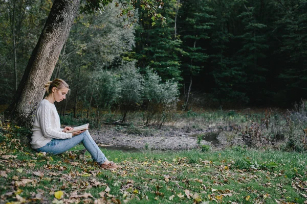 Jovem mulher em suéter branco e jeans azul lê um livro sob uma árvore no parque — Fotografia de Stock