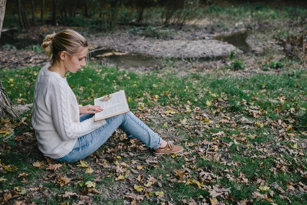 Jovem mulher em camisola branca e jeans azul lê um livro no parque — Fotografia de Stock