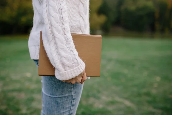 Przycięte zdjęcie młodej kobiety w biały sweter i dżinsy, trzymając w ręku książkę na zewnątrz w parku — Zdjęcie stockowe