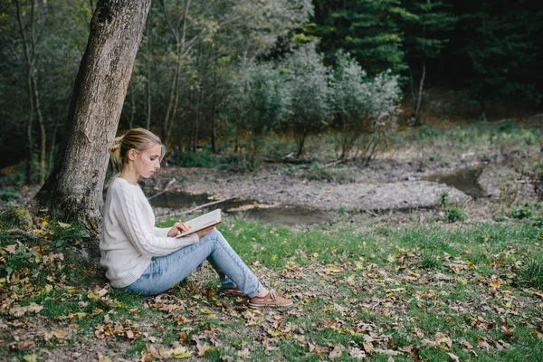 Jovem mulher em suéter branco e jeans azul lê um livro sob uma árvore no parque — Fotografia de Stock