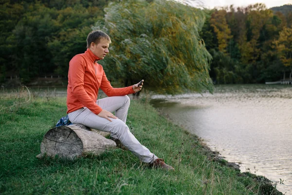 Joven en un suéter rojo relajante al aire libre cerca del lago y utiliza un teléfono móvil — Foto de Stock