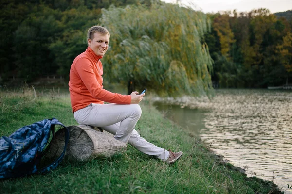 Młody człowiek w czerwonym swetrze relaks na świeżym powietrzu w pobliżu jeziora i używa telefonu komórkowego — Zdjęcie stockowe