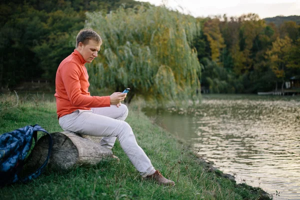 Joven en un suéter rojo relajante al aire libre cerca del lago y utiliza un teléfono móvil — Foto de Stock