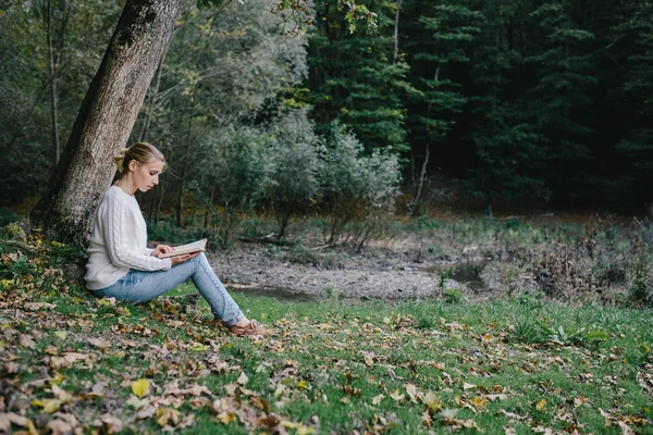 Jovem mulher em suéter branco e jeans azul lê um livro sob uma árvore no parque — Fotografia de Stock